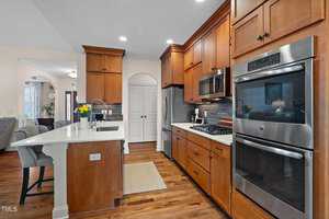 Modern kitchen with wooden cabinets, stainless steel appliances, and a double wall oven. A large island features a sink and barstools. The room has hardwood flooring and recessed lighting, with a partial view into a living area with a dining table.