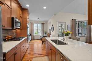 A bright kitchen with wooden cabinets, a white marble island with a sink, and stainless steel appliances. In the background, there's a cozy seating area with a chair and table near large windows with beige curtains, letting in natural light.