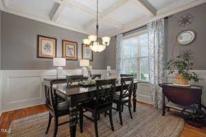 A dining room with a dark wood table and six chairs, a chandelier above, and a sideboard with two lamps. The walls are gray with white wainscoting, decorated with framed art. A window with patterned curtains and natural light brightens the space.