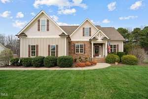A charming single-story house with a mix of stone and siding exterior. The house features multiple gables, a well-maintained lawn, shrubs, and a U.S. flag by the door. The sky is blue with scattered clouds.