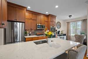 A modern kitchen with wooden cabinets, stainless steel appliances, and a marble island with a sink. A vase with colorful flowers sits on the island. There's a large window with curtains and a cozy chair nearby.