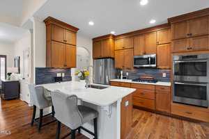 Modern kitchen with wooden cabinets, white countertops, and stainless steel appliances, including a double oven and microwave. Gray upholstered barstools line the island. Recessed lighting and a vase with flowers add a cozy touch.