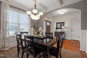 A dining room with a dark wooden table and six chairs. The table has three candles and decorative items. A modern chandelier hangs above. Light streams in through a window with patterned curtains. The room has hardwood floors and an entryway in the background.