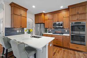 A modern kitchen with wooden cabinets, stainless steel appliances, and a marble island with two cushioned chairs. The backsplash is gray with a brick pattern, and a vase with flowers decorates the island. Warm lighting brightens the space.