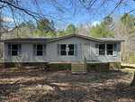 An off-white mobile home at 4131 Doe Court features a small wooden front porch set amidst the wooded surroundings of Oxford. The ground is covered with dry leaves, and trees with bare branches encircle the home, while the sky remains partly cloudy.