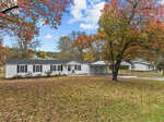 A white, single-story house with dark shutters on 2204a Sam Moss Hayes Road is surrounded by autumn trees. A carport is attached to the right side, and fallen leaves cover the grass in the spacious Creedmoor yard under a partly cloudy sky.