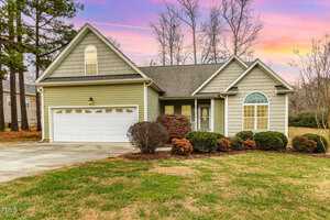 A single-story house with a gable roof, double garage, and a front yard with shrubs. The home exterior is light brown with white trim. The sky in the background has a colorful sunset with hues of orange and pink.