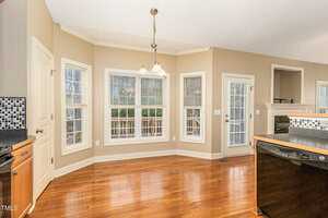 A bright kitchen with wooden floors, beige walls, and large windows. It features black and white kitchen counters, a dishwasher, and a hanging light fixture above the breakfast area. There's a view of trees outside the windows.