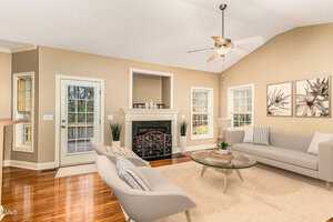 A spacious living room with high ceilings features a modern fireplace, tan walls, and wooden floors. Two beige sofas face a glass coffee table, and large windows allow natural light to fill the room. A ceiling fan is centered above.