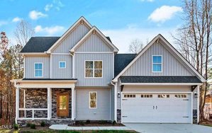 The two-story house at 1765 River Club Way in Franklinton features light gray siding, white trim, and a dark shingled roof. It boasts a wooden front door, a covered porch with stone accents, and a two-car garage. The scenic background is filled with trees against a blue sky topped with clouds.