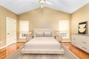 A bright bedroom with vaulted ceiling, featuring a large bed with beige linens, two bedside tables, and a dresser. Soft lighting from the windows adds warmth to the neutral walls and polished wooden floor. A decorative piece hangs above the dresser.