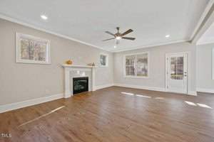 A spacious living room with wood flooring, a white fireplace, and a ceiling fan. Two windows and a glass door let in natural light. Walls are painted light gray, and there are autumn decorations on the mantel.
