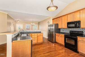 A spacious kitchen with wooden cabinets, dark countertops, and stainless steel appliances, including a fridge and oven. The room features hardwood floors, a pendant light, and a mosaic tile backsplash. A breakfast bar separates the open space.
