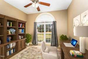 A home office with a large arched window allowing natural light in. It features a wooden bookcase filled with books, a desk with a laptop, and a cushioned chair. Decor includes a ceiling fan, potted plants, and neutral-colored walls.