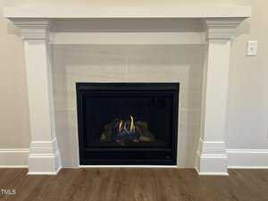 A fireplace with a lit gas fire is set in a white mantle with decorative columns. The hearth features a black frame around the flames, which dance among realistic logs. The flooring in front is hardwood.