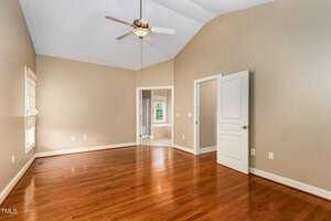 An empty room with light brown walls and polished wooden floors. There's a ceiling fan above, two windows on the left, and an open door leading to a bathroom. The room is well-lit with natural light.