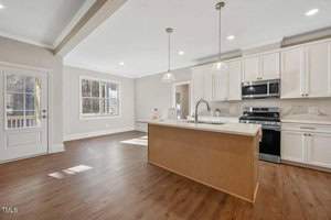 Modern kitchen with hardwood floors, a large island, white cabinets, and stainless steel appliances. Two pendant lights hang above the island. Natural light streams in through a window and glass door.