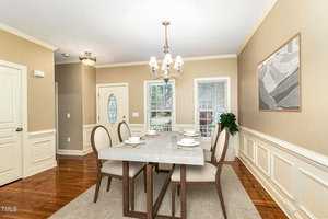 A dining room with a large rectangular table set for six, featuring cream-colored plates and glasses. The room has beige walls, wood flooring, a chandelier, a painting on the wall, and windows with white trim, letting in natural light.