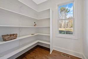 A small pantry with white shelves, featuring a basket, a bottle, and some envelopes. There's a large window providing natural light, and a view of trees outside. The room has wooden flooring.