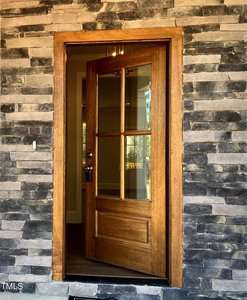 A wooden door with glass panes is slightly open, set in a stone wall with a mix of gray and brown bricks. Natural light streams through the glass, illuminating the interior.