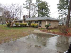 A single-story brick house with a gray roof is surrounded by wet ground, trees, and shrubs. A concrete driveway curves towards the front entrance. The sky appears overcast, suggesting recent rain.