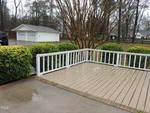 A rain-soaked wooden deck with white railings overlooks a driveway and a detached two-car garage. Wet pavement and trimmed hedges frame the scene, with overcast skies and bare trees in the background.