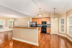 A modern kitchen with light wooden cabinets, black appliances, and a central island with a wooden countertop. The kitchen is adjacent to an open-plan dining area with large windows and hardwood floors. Two pendant lights hang above the island.