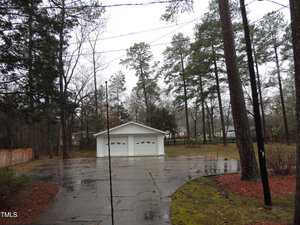 A small white garage is situated at the end of a curved driveway, surrounded by tall pine trees. The ground is wet from rain, and the sky is overcast, creating a damp, serene atmosphere. A house is partially visible in the background.