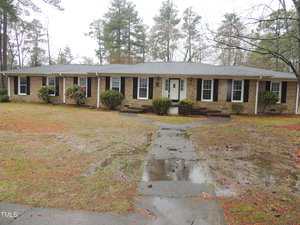 A single-story brick house with white trim and multiple windows, surrounded by a yard with some shrubs and trees. The ground is wet, with puddles visible on the sidewalk leading to the entrance. Overcast sky overhead.
