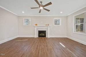 A spacious, empty living room with light wooden floors and a ceiling fan. The room features a white fireplace with a small pumpkin and decor items. Two windows flank the fireplace, and another window is on the right wall.