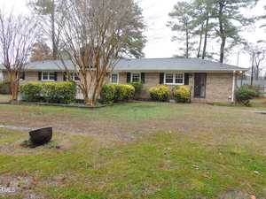 A one-story brick house with a gray roof is surrounded by bare trees and yellow shrubs. The front yard is grassy with scattered dry leaves, and a black object rests on the lawn. The sky is overcast, suggesting a chilly day.