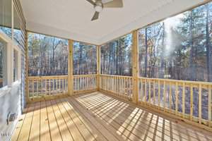 A sunlit wooden screened porch with a ceiling fan overlooks a forested area. The sun casts shadows of the railings onto the wooden floor, creating a striped pattern. Trees are visible through the screened walls.