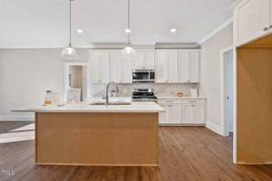 A modern kitchen with a central island featuring a sink and faucet. The kitchen includes white cabinets, a stainless steel oven, and a microwave. Pendant lights hang above the island, and the floor is finished with brown wooden planks.