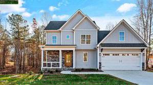 A modern two-story house with gray siding, a front porch, and a double garage. It is surrounded by leafless trees and a grassy lawn, set against a clear blue sky.