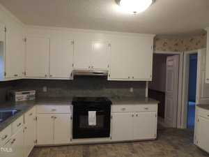 A kitchen with white cabinets, a black electric stove, and gray countertops. A floral wallpaper decorates the walls. The floor is tiled in a neutral tone, and a small window is visible above the sink.