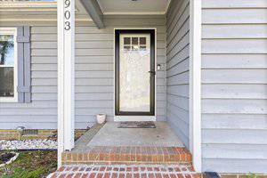 A gray house entryway with a glass storm door featuring a "Welcome" mat. The door is flanked by light and dark gray siding, and there's a small white planter on the porch. The house number "903" is visible on a vertical post.