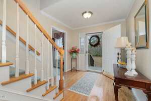 A welcoming foyer with wooden stairs, a small table holding a lamp and a bowl of lemons, and an open door adorned with a wreath. A floral arrangement stands on a pedestal. Light wooden flooring and beige walls complete the cozy look.