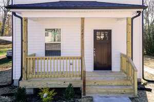 Front view of a house with a small wooden porch, stairs, and railing. The door is dark-colored, and the exterior walls are white. A window with closed blinds is to the left of the door. Small plants are in front of the porch.