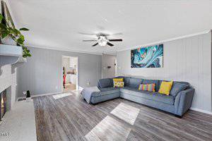 A modern living room with a gray sectional sofa and colorful pillows. A ceiling fan is overhead, and a painting hangs on the wall. The room features light wood flooring and white walls. Sunlight streams through a window, and a doorway leads to a kitchen.