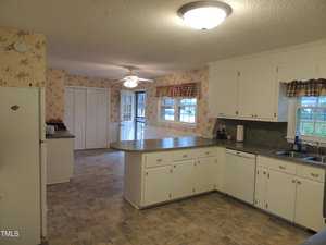 A spacious kitchen with white cabinets and a large island. There's a ceiling fan and large windows with checkered valances. The floor is tiled, and the walls are adorned with floral wallpaper. A door leads to an adjoining room.