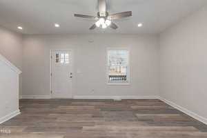 An empty living room with light gray walls and wood-look flooring. A ceiling fan with lights is mounted on the ceiling. The room features a door and a window with blinds, and there's a staircase on the left side.