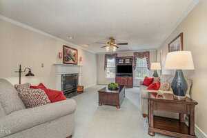 A cozy living room with beige walls features a fireplace, two gray sofas with red cushions, a wooden coffee table, and a TV on a stand. A ceiling fan hangs above, and a large window lets in natural light. Decor includes lamps and framed art.
