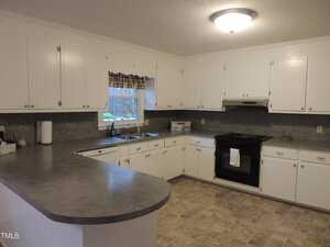 A spacious kitchen with white cabinets, a dark countertop, and a black stove. The room has tiled flooring and a window with a plaid valance above the sink, allowing natural light in. A ceiling light fixture provides additional illumination.