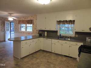 A spacious kitchen with white cabinets and a gray countertop. An island divides the room, with large windows letting in natural light. The floor is tiled, and floral wallpaper decorates the walls. A dining area with a ceiling fan is visible in the background.