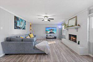 Spacious living room with a gray sectional sofa and yellow pillows. A ceiling fan is above the sofa. The room features a wall-mounted TV showing a beach scene, a white brick fireplace, and a large mirror. Decor includes a painting and a potted plant.