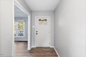 A small, bright entryway with a white front door, a small glass panel above the door, and light gray walls. The floor is wooden, and a doorway on the left leads to a room with large windows and a view of trees outside.