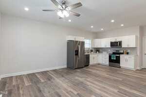 A modern kitchen with white cabinets, stainless steel appliances including a fridge and oven. It features wooden flooring, a ceiling fan with lights, and an uncluttered countertop near a window. The room is spacious and well-lit.