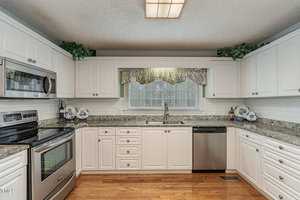 A bright kitchen with white cabinets, stainless steel appliances, and a granite countertop. The window above the sink features a floral valance. Decorative plates adorn the countertop corners, and potted plants are on the cabinet tops.