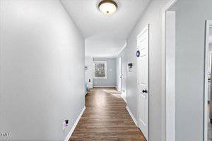A hallway with light gray walls and wooden flooring leads to a room with a large window. On the left, a wall-mounted light fixture is seen, and on the right, there are closed white doors with black handles. Natural light brightens the space.