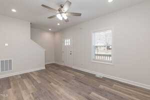 A bright, empty room with light gray walls and wood flooring. A ceiling fan with lights hangs from the ceiling. A window with blinds is on the right, and a door with a small window is on the wall opposite the stairs.